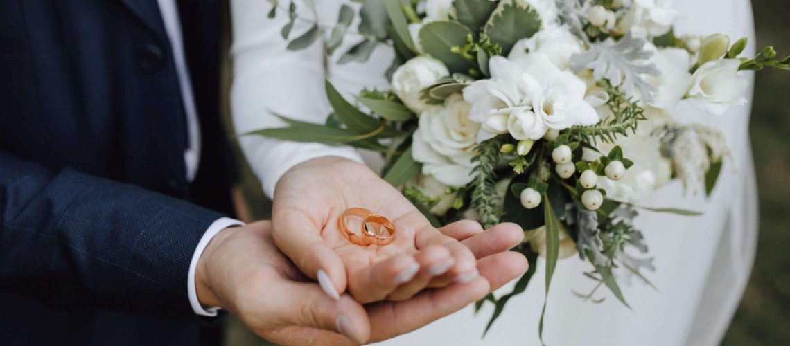 Wedding bands in the hands of bride and groom and with beautiful wedding bouquet made of greenery and white flowers